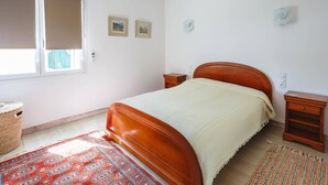 Un dormitorio con cama de madera, una ventana con persianas, dos mesitas de noche, una cesta tejida y una alfombra estampada.