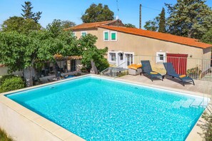 Une piscine avec des chaises longues, un bĂątiment dotĂ© dâun toit en tuiles et des arbres en arriĂšre-plan.