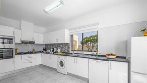 Une cuisine moderne avec des meubles blancs, un four encastré, un lave-linge et une grande fenêtre donnant sur un arbre.