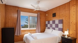 A bedroom with a bed, a nightstand, a ceiling fan, a window with curtains, and a view of the outside. - Villa Casa Jaska by Villa Plus (Urbanización Riviera Sol)