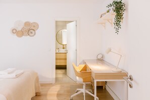 A minimalist bedroom with a wooden desk, a chair, a bed, and a bathroom visible through an open door.