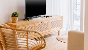 A modern living room with a flat-screen TV on a wooden console, a wicker chair, a small potted plant, and a white curtain. - FLH Caparica By the Sea with Balcony (Almada)