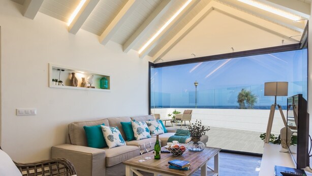 A modern living room with a large glass window overlooking a beach, a sofa with teal cushions, and a coffee table with books and a bottle. - Villa Del Mar by Villa Plus (Caleta de Fustes)