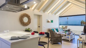 A modern kitchen with a central island, a dining area with a sofa and coffee table, and a large window overlooking the sea. - Villa Del Mar by Villa Plus (Caleta de Fustes)