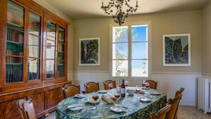 Une salle à manger avec un grand buffet en bois, une table dressée pour six personnes avec des verres à vin, des assiettes et une bouteille. Il y a deux fenêtres ornées de tableaux, un lustre et un radiateur.