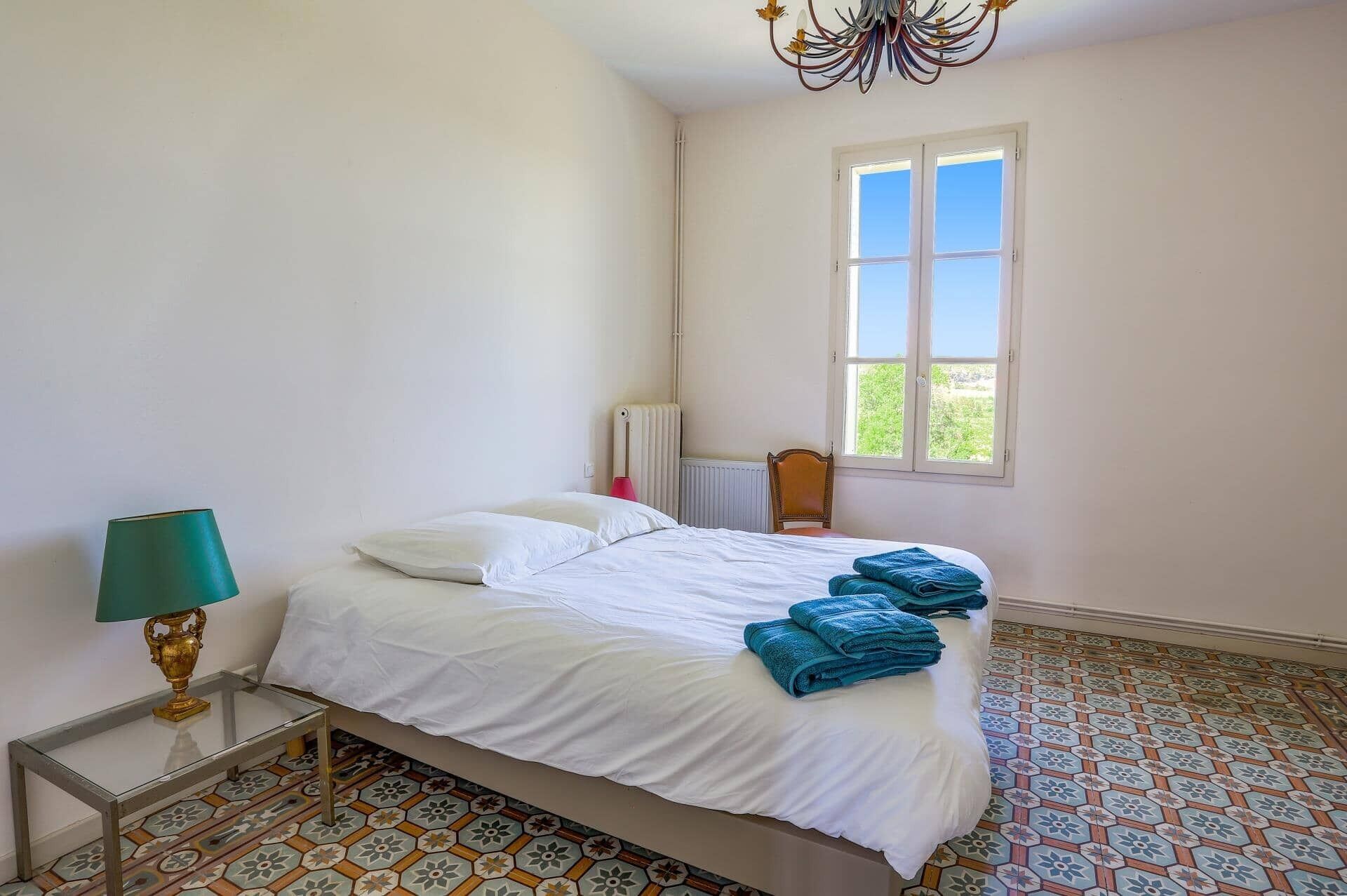 A bedroom with a bed, a radiator, a window with a view, and patterned floor tiles.