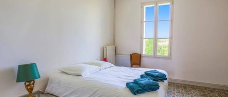 A bedroom with a bed, a radiator, a window with a view, and patterned floor tiles.