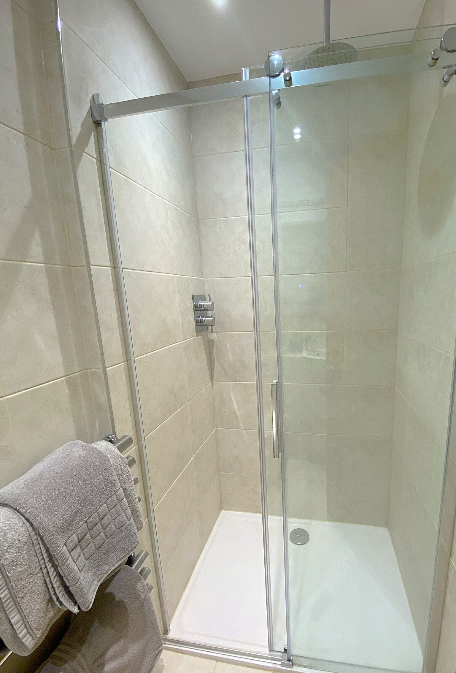 A glass-enclosed shower with a white floor, a towel rack with a folded towel, and a visible control panel on the wall.