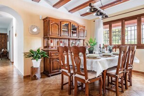 A dining area with a wooden table set for six, surrounded by six wooden chairs. A large wooden cabinet with glass doors is against the wall. - Villa Estiu by Villa Plus (Jávea)