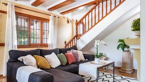 Una sala de estar con un sofá oscuro, varios cojines, una mesita auxiliar, una planta en maceta y una escalera.