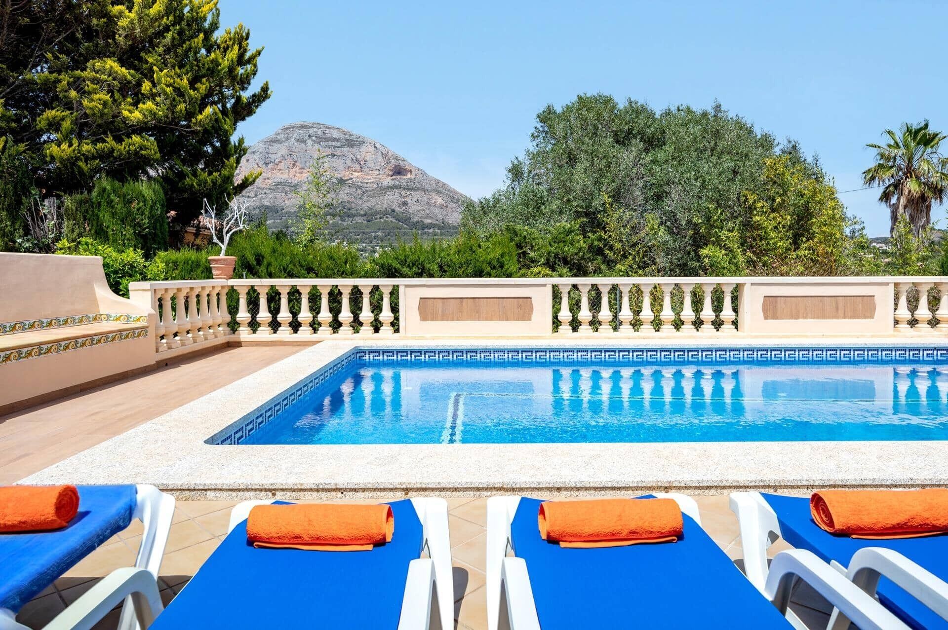 Ein Pool mit blauen Fliesen, umgeben von Liegestühlen mit orangefarbenen Handtüchern, einem dekorativen Geländer und einem Berg im Hintergrund.