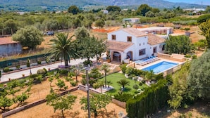Una villa blanca con piscina, rodeada de jardines ajardinados y palmeras, con montañas al fondo.