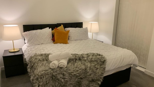 A neatly made bed with a fluffy gray throw, white bedding, and a single yellow pillow. There are two bedside tables with lamps, and a window with curtains. - Pristine 3-bed House in Grays (Grays)