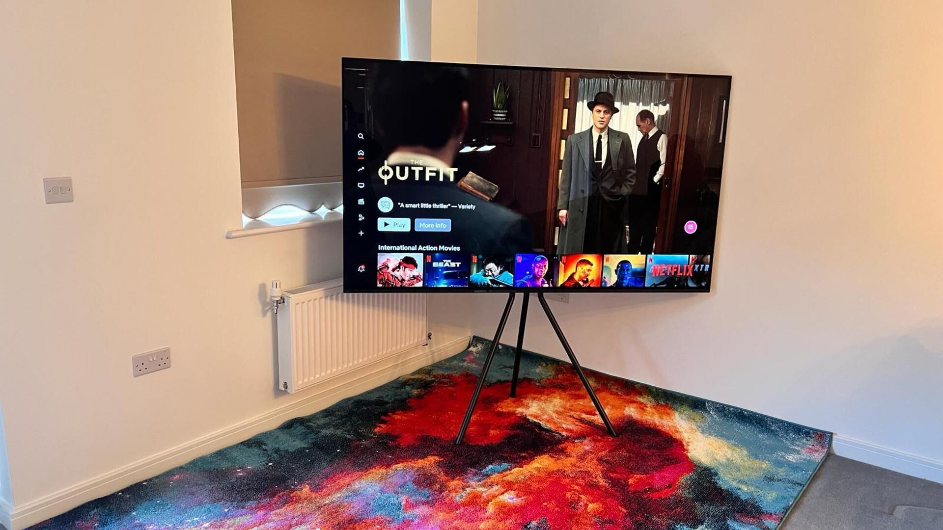 A flat-screen TV on a stand with a colorful abstract floor design, a shelf above it, and a wall-mounted thermostat.