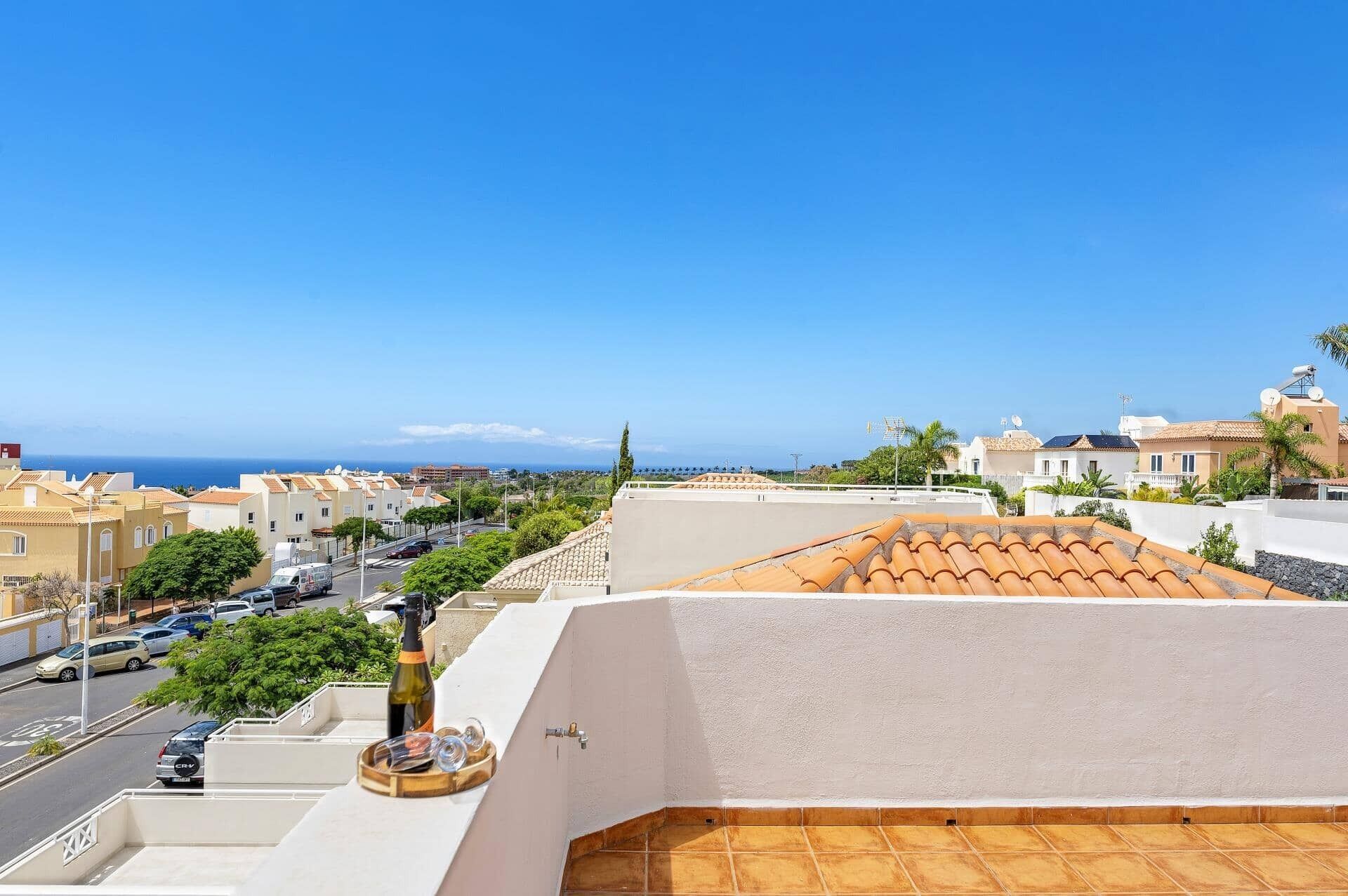 A rooftop terrace with a view of the sea, a street with parked cars, and buildings with Mediterranean architecture.