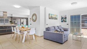 Un salon moderne avec un canapé gris, des chaises blanches et un coin repas comprenant une table et des chaises en bois. On aperçoit en arrière-plan une cuisine équipée d’appareils électroménagers en acier inoxydable.
