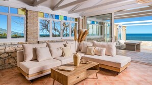A spacious living area with a stone wall, a large sofa, a wooden coffee table, and a view of the sea through large windows. - Villa Ionio Beach House by Villa Plus (Ano Vasilikos)