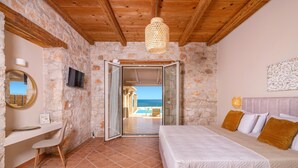 A bedroom with a stone wall, wooden ceiling, a bed with pillows, a desk with a chair, and a TV. - Villa Ionio Beach House by Villa Plus (Ano Vasilikos)
