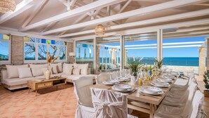 A dining area with a view of the sea, a sofa, and a table set for a meal. - Villa Ionio Beach House by Villa Plus (Ano Vasilikos)