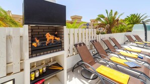 Un barbecue, avec un téléviseur à écran plat placé juste au-dessus, entouré de fauteuils de jardin dotés de coussins jaunes.