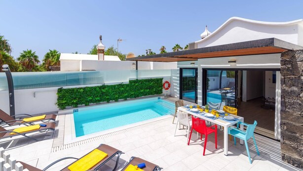Un espace piscine extérieur moderne, doté d’une table et de chaises, et entouré d’un bâtiment blanc à la façade en pierre.