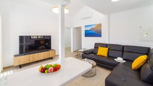 Un salon moderne avec un canapé en cuir noir, un meuble en bois, un téléviseur à écran plat et un panier de fruits posé sur une table blanche.