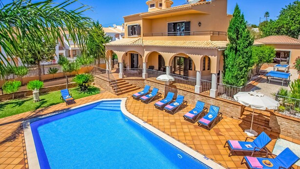 A poolside area with sun loungers, a swimming pool, and a building with a tiled roof and arches. - Villa Karlia by Villa Plus (Vale Rebelho)