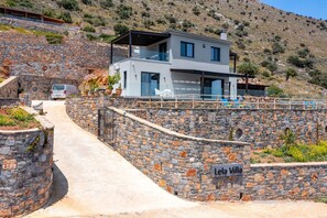 Una villa moderna con entrada de muro de piedra, balcón con mobiliario exterior y vistas a un paisaje montañoso.