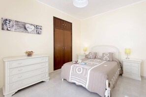 A bedroom with a bed, two bedside tables, a dresser, a wardrobe, and a wall with framed pictures.