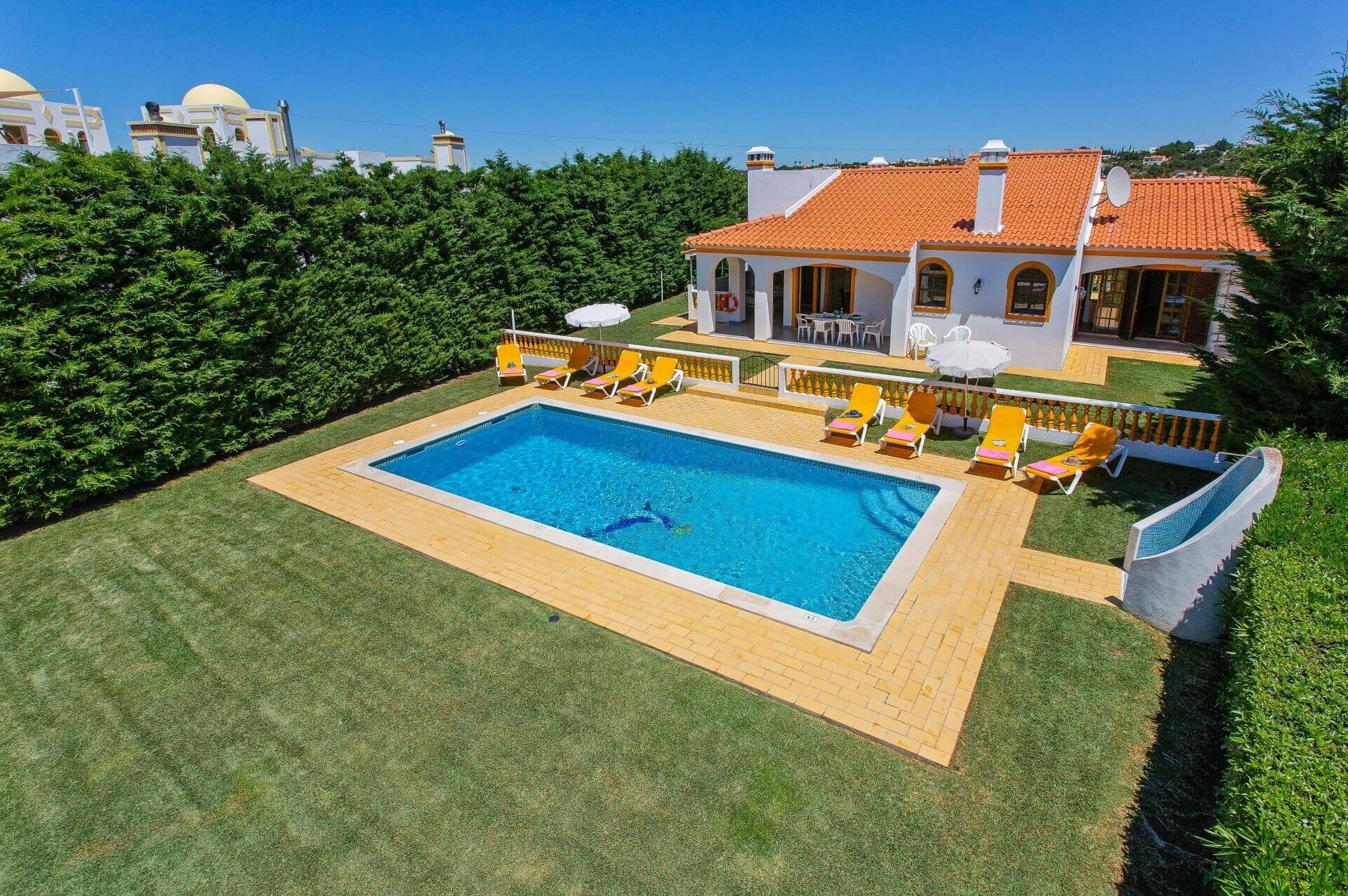 A large rectangular swimming pool with a tiled deck, surrounded by a well-maintained lawn and a white building with a red-tiled roof.