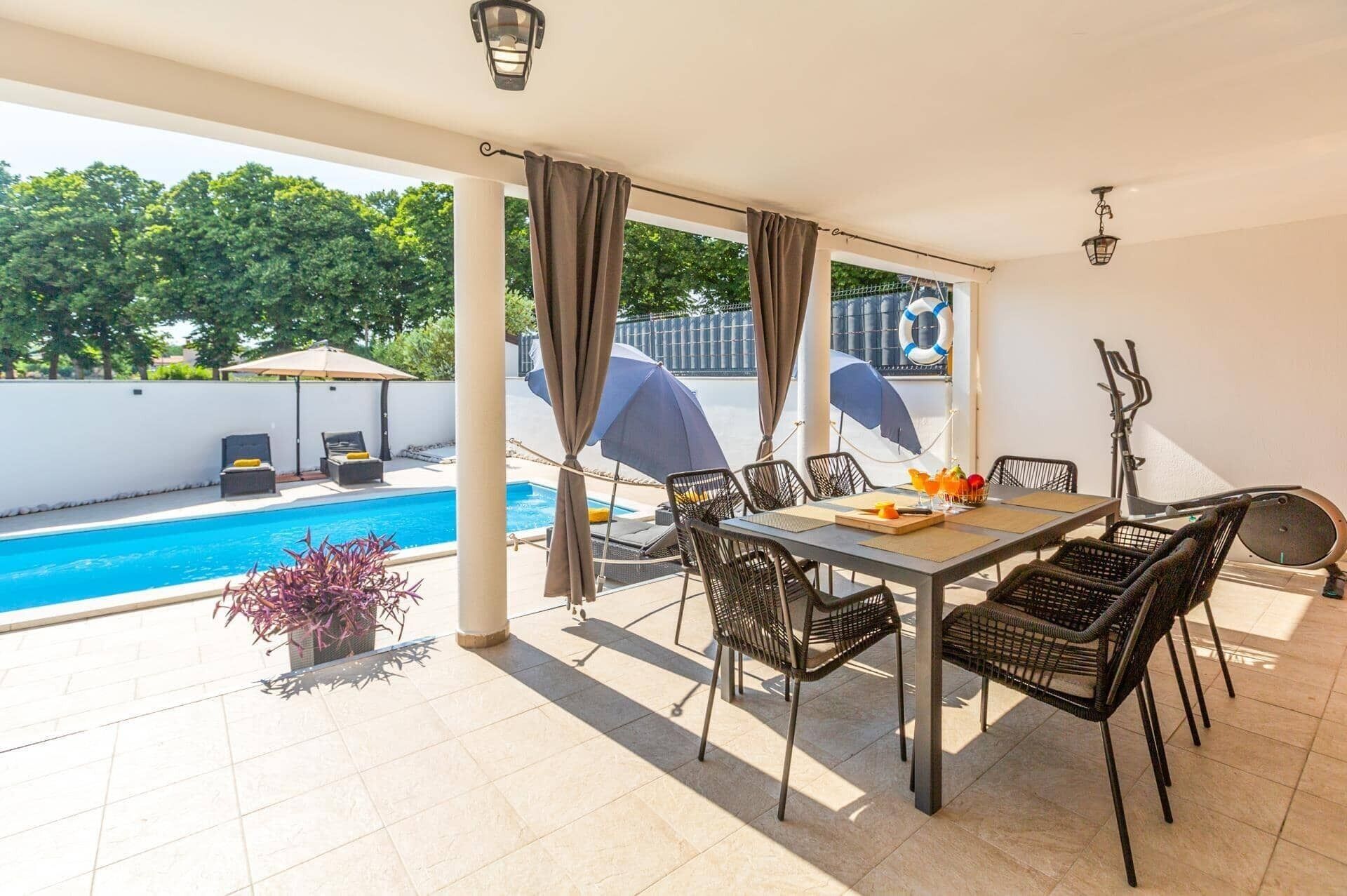A covered outdoor dining area with a pool view, featuring a table and chairs, umbrellas, and a lifebuoy on the wall.
