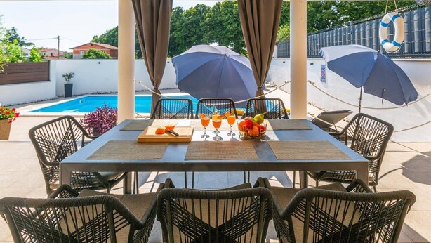 An outdoor dining area with a pool view, featuring a table set with drinks and fruits, surrounded by wicker chairs. - Villa Melani Vita by Villa Plus (Marčana)