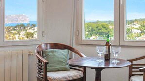 A room with a view of a coastal landscape, a wooden chair with a cushion, a small round table with a bottle and glasses, and a radiator. - Villa Murales by Villa Plus (El Tosalet)