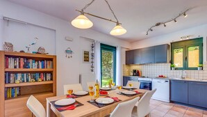 A modern kitchen with a dining area, featuring a wooden table set for four, a bookshelf, and a view of the outdoors. - Villa Nefeli Green by Villa Plus (Chersonisos)