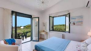 A bedroom with a bed, a desk, and a chair. There are two windows with open shutters, and a balcony with a table and chairs. - Villa Nefeli Green by Villa Plus (Chersonisos)
