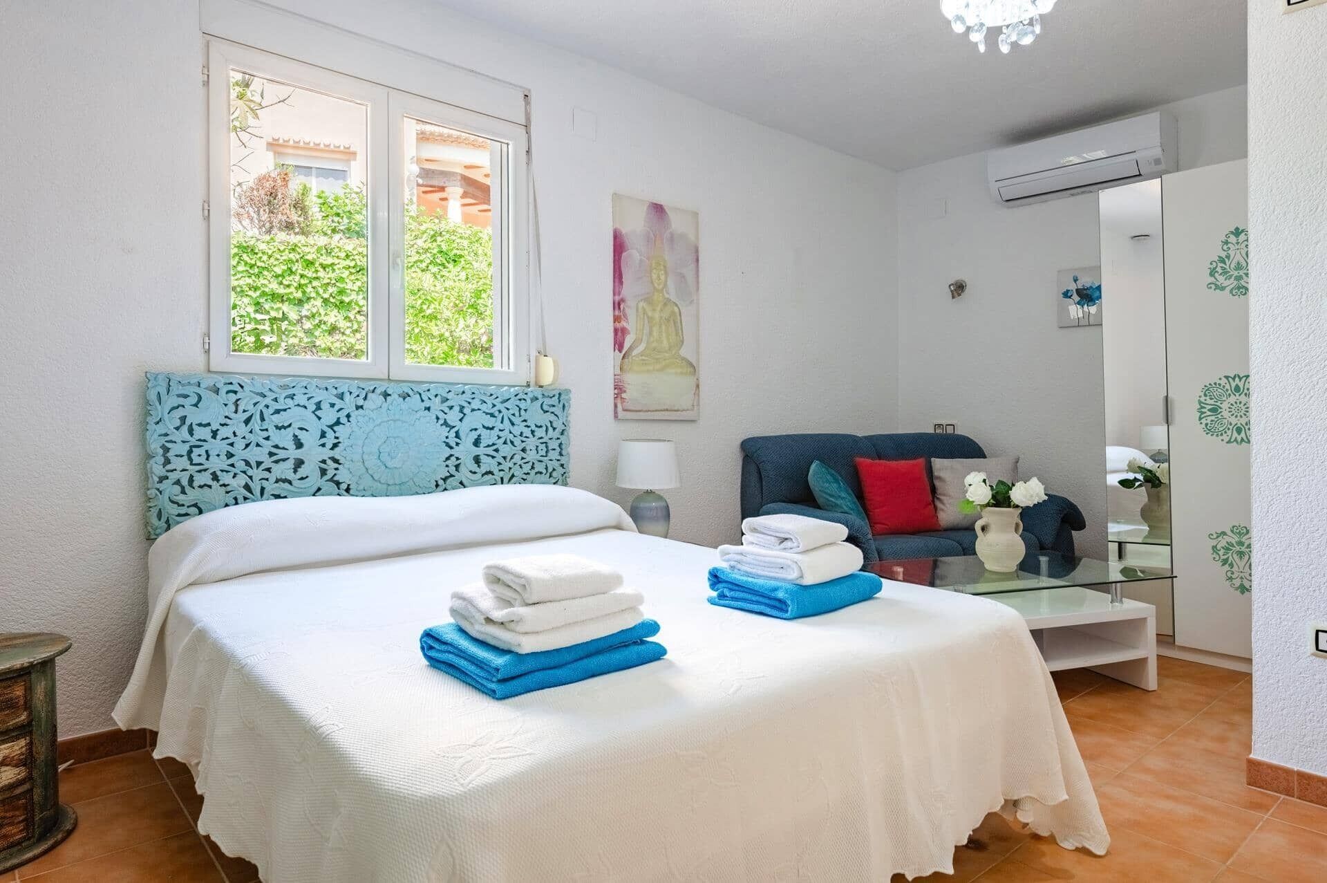 A bedroom with a bed, a sofa, a glass table with a vase, and a window with a view of greenery.