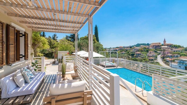 A balcony with a pool, wooden furniture, and a view of a town with a church. - Villa Observer by Villa Plus (Ionian Islands)