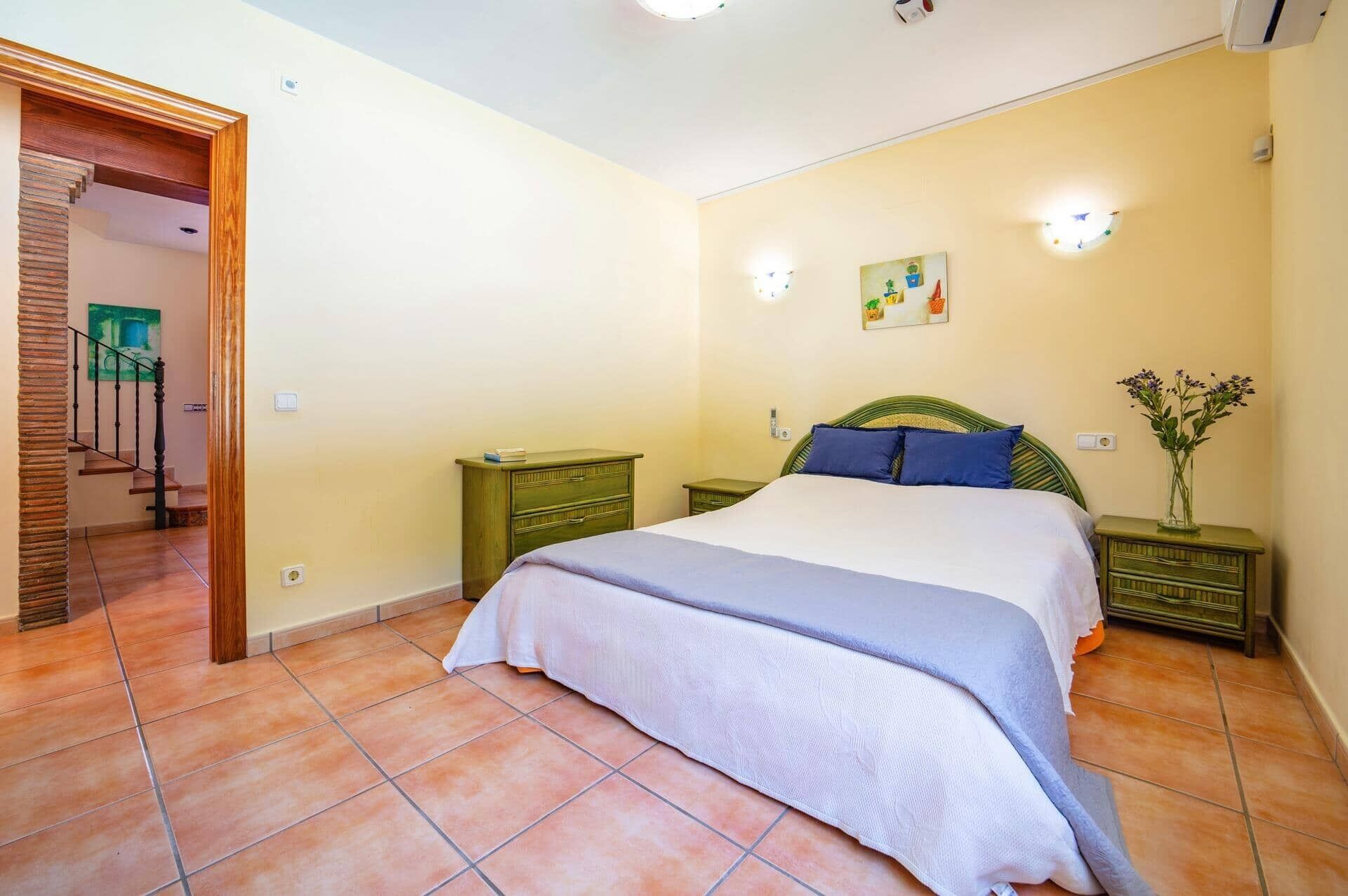 Ein Schlafzimmer mit Bett, Nachttischen, einem Schminktisch und einem Gemälde. Eine Treppe führt in einen anderen Raum.