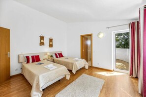 A room with two beds, a wooden floor, a white wall, a door, a window with curtains, and a small table with a lamp.