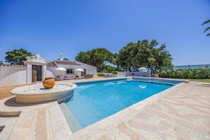 En swimmingpool med fliseterrasse, en hvid bygning med fliset tag og et stort træ i baggrunden.