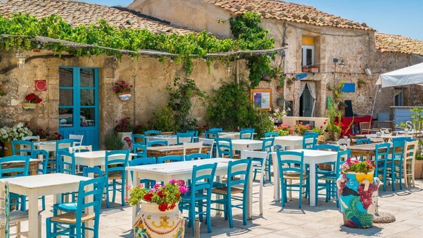Un café al aire libre con mesas blancas y sillas azules, rodeado de edificios de piedra con tejados de tejas y vegetación.