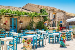 An outdoor café with blue chairs and tables, surrounded by a stone building with a tiled roof and greenery.