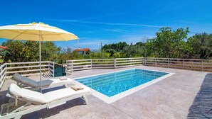 Ein rechteckiger Swimmingpool mit weißer Liegefläche, zwei Liegestühlen und einem gelben Sonnenschirm.