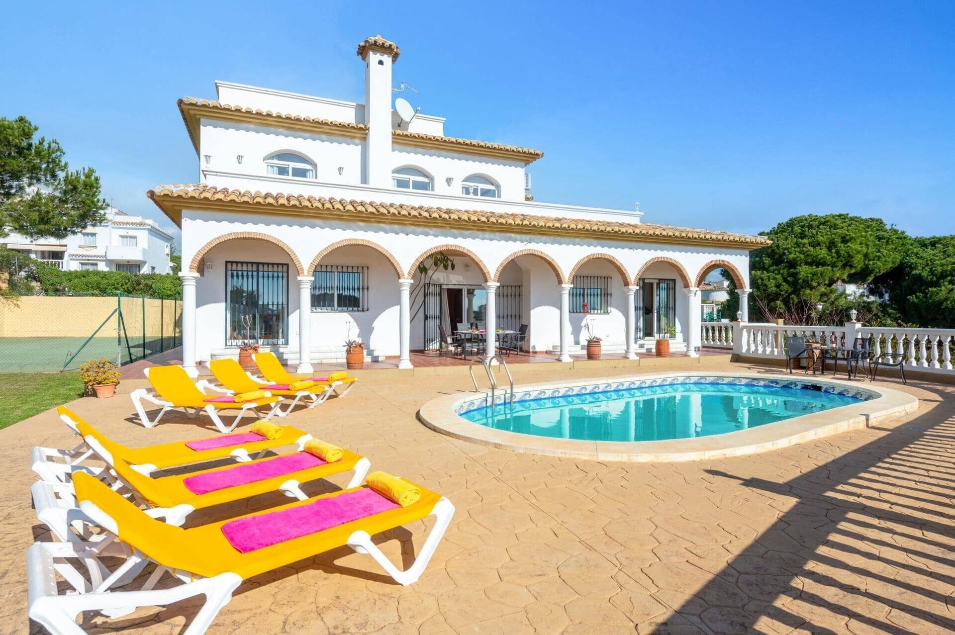 A white villa with a pool, yellow lounge chairs, and a covered patio area.