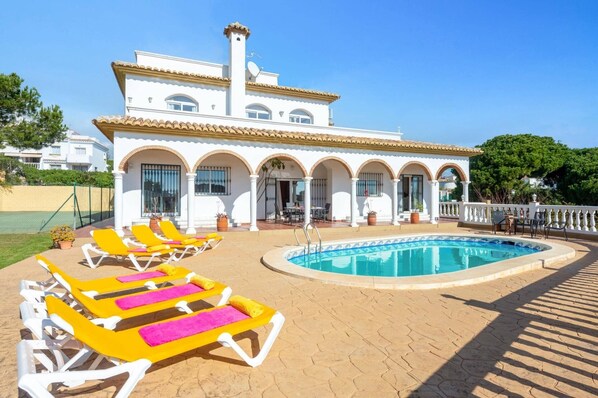 A white villa with a pool, yellow lounge chairs, and a covered patio area.