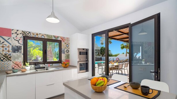 A modern kitchen with a large window, a dining area with a pool view, and a built-in oven. - Villa Taiga by Villa Plus (Playa Blanca)