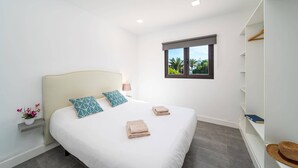 A bedroom with a bed, pillows, a window, a bedside table with a lamp, a plant, and a shelf with a book and a hat. - Villa Taiga by Villa Plus (Playa Blanca)
