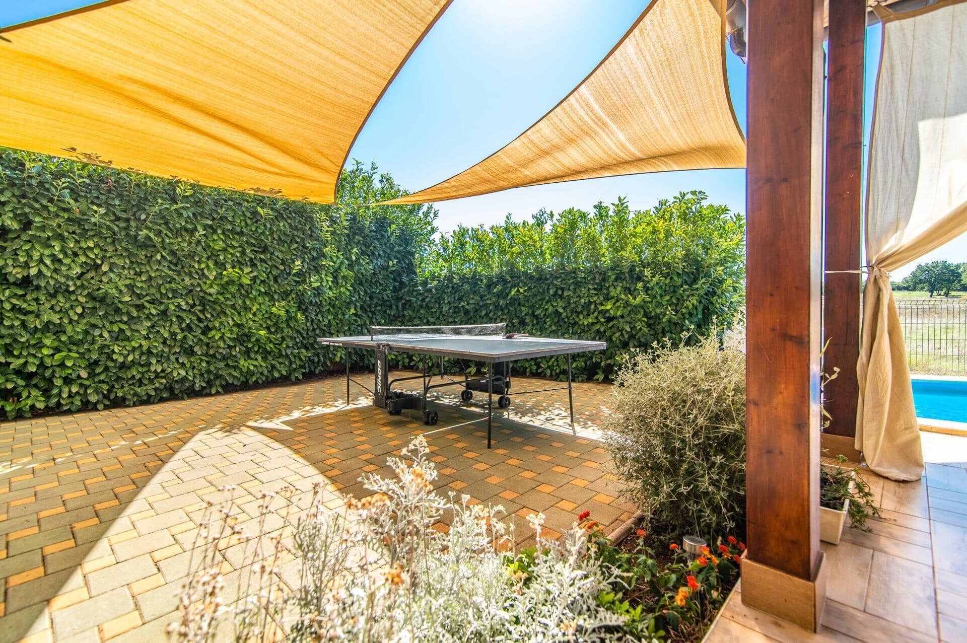 Ein Ping Pong Tisch steht in einem überdachten Außenbereich mit einer Heckenwand, Ziegelböden und einem Pool im Hintergrund.