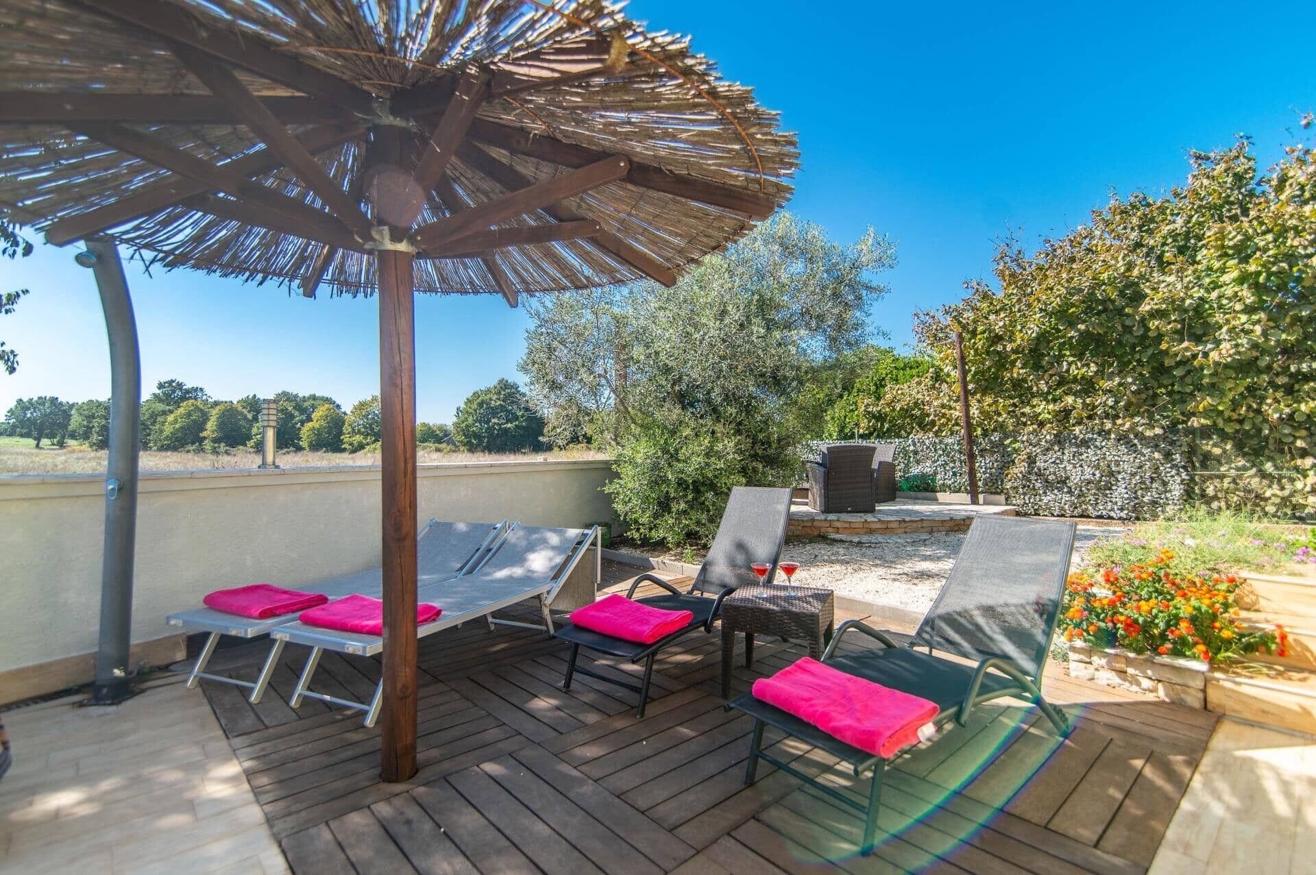 Patio esterno con pavimento in legno, poltrone con cuscini rosa, ombrellone di paglia e vista su alberi e cielo sereno.
