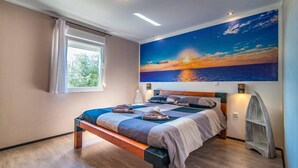 Une chambre à coucher avec un lit, une fenêtre, un tableau représentant un coucher de soleil et un banc en bois.